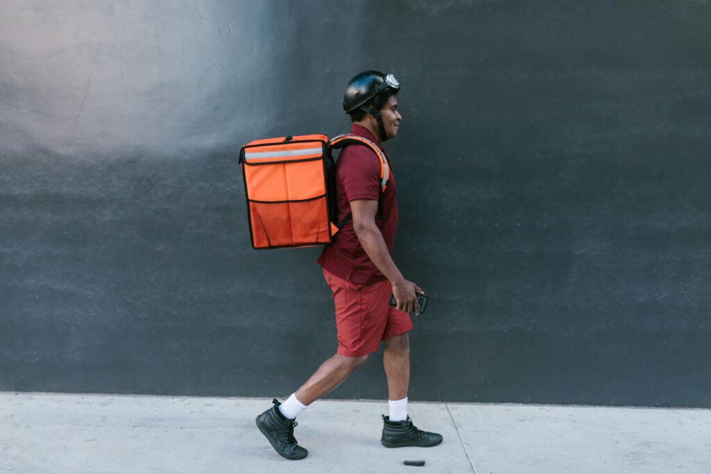 A delivery person in red uniform and helmet walks with an insulated bag against a city backdrop.