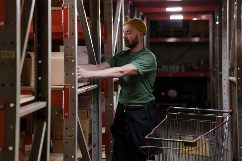 Male warehouse worker in a beanie organizes inventory on shelves with a push cart indoors.