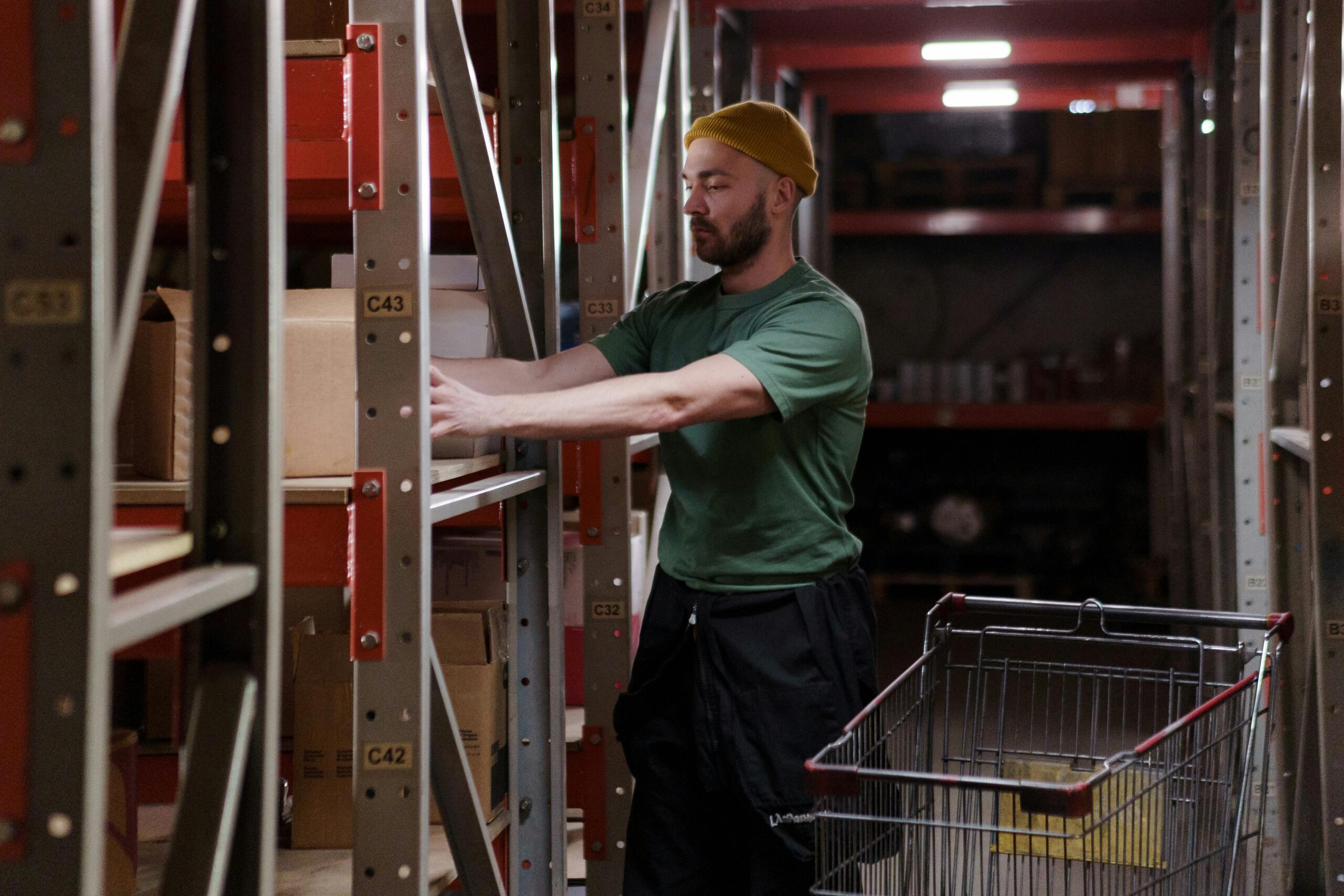 Male warehouse worker in a beanie organizes inventory on shelves with a push cart indoors.